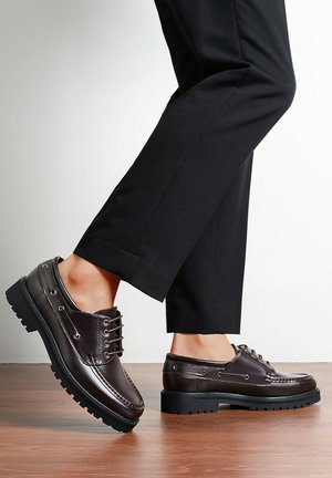 Personne portant un pantalon noir et des chaussures en cuir marron foncé à lacets, debout sur un sol en bois devant un mur blanc.