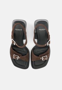 Ζευγάρι μαύρων σανδαλιών με καφέ σουέντ λουριά και ασημένιες αγκράφες, τετράγωνες μύτες και το λογότυπο "Vagabond Shoemakers" στις σόλες.