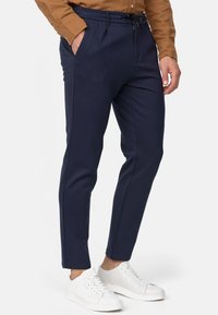 Pantalones tapered en color azul marino, confeccionados en tejido suave, con cordón en la cintura y bolsillos laterales, combinados con zapatillas blancas.