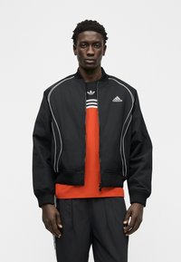 Άνδρας που φοράει μαύρο μπουφάν Adidas bomber πάνω από κόκκινη μπλούζα με λογότυπα Adidas, συνδυασμένο με μαύρο παντελόνι φόρμας Adidas, σε απλό φόντο.
