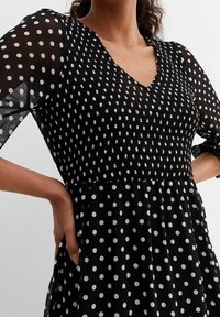 Schwarzes Kleid mit weißen Punkten, ausgestattet mit einem V-Ausschnitt, geraffter Taille und durchsichtigen Puffärmeln. Der Stoff hat ein strukturiertes Aussehen.