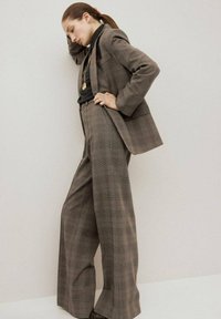 Kobieta w brązowym garniturze w kratę z szerokimi spodniami i marynarką, czarnym topem i złotym naszyjnikiem, pozująca z ręką na głowie na tle jednolitej ściany.