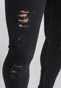 Mustat farkut, joissa on kapea malli ja kuluneita repeämiä polven alueella, rispaantuneet reunat ja sileä pinta.