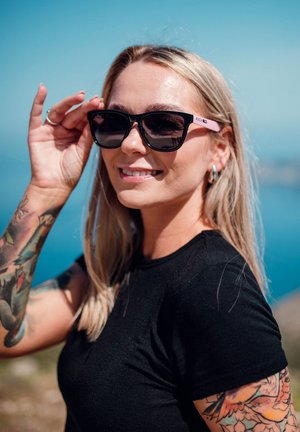 Černé sluneční brýle s silným rámem a růžovými akcenty, které nosí osoba, jež si je upravuje, s světlými hnědými vlasy a viditelnými barevnými tetováními.