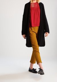 Schwarze gestrickte Strickjacke, rotes plissiertes Top mit Spitzenakzenten, senfgelbe schmale Hose und schwarze Schuhe mit Schleifen und strukturierten Sohlen.