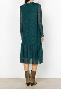 Vestido verde azulado con mangas largas y transparentes, con un patrón de hojas y un dobladillo volado. Combinado con botas de tobillo marrones.