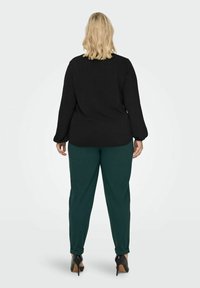 Woman with shoulder-length blonde hair wearing a black long-sleeve top, dark green pants, and black high-heeled shoes standing facing away.