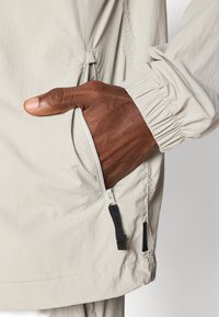 Giacca leggera beige chiaro con polsino elastico, tasca laterale con chiusura a zip e design minimalista. Texture liscia, leggermente stropicciata.