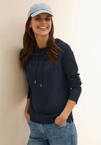 Mörkblå huvtröja med dragsko och logotyp på framsidan, kombinerad med en ljusblå keps. Modellen har på sig ljusa jeans, med händerna i fickorna.