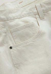 Gros plan sur la poche arrière d'un jean en denim couleur crème avec passant de ceinture, détail rivet et étiquette de marque "JORT" visible à l’intérieur de la ceinture.