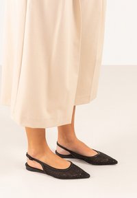 Los zapatos planos negros tipo slingback tienen una punta afilada, diseño de malla y correa ajustable en el tobillo. Se llevan con unos pantalones de pierna ancha de un beige claro.