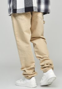 Pantaloni beige larghi con una texture liscia, dotati di due tasche posteriori, abbinati a scarpe da ginnastica bianche e una camicia a quadri bianco e nero.
