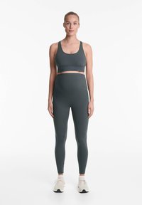 Sötétzöld sportmelltartó és magas derekú leggings rugalmas anyagból, világos színű sportcipővel párosítva.