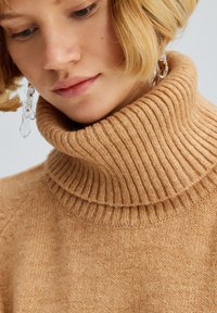 Beige, ribbet rullekravesweater med en blød tekstur; har en bred høj krave, der fremhæver et jævnt strikkemønster og en varm farvetone.