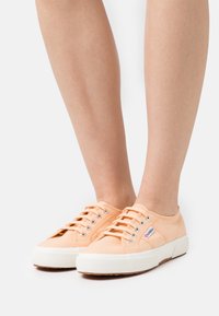 Broskvové plátěné tenisky s bílými pryžovými podrážkami a průchodkami, mají pět tkaniček a malý logo štítek na boku.