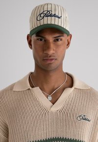 Polo en tricot beige avec un col en V et des rayures vertes ; associé à une casquette rayée beige avec des broderies bleu marine et une visière verte.