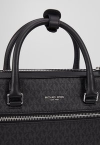 Sac à main noir Michael Kors avec doubles poignées, fermeture éclair argentée et texte du logo "MICHAEL KORS EST 1981" sur une surface à motifs.
