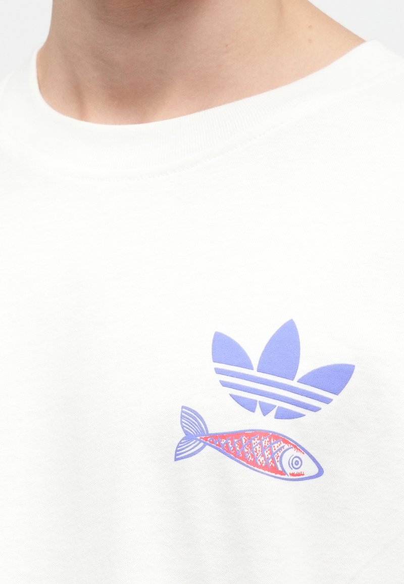 White cotton t-shirt featuring a purple adidas logo above a stylized fish in blue and red decorative patterns.