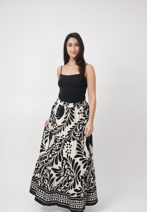 Femme debout sur un fond blanc portant un haut noir sans manches et une longue jupe blanche avec des motifs floraux noirs audacieux.