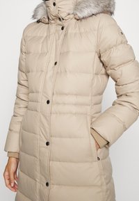 Beige pufferjack met een met bont afgewerkte capuchon, voorzien van een gewatteerd patroon, drukknoopsluiting en zijzakken. Zacht, geïsoleerd materiaal.