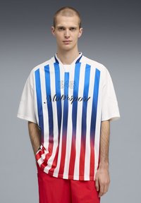 Νεαρός με κοντά μαλλιά, ντυμένος με λευκό πουκάμισο με μπλε και κόκκινες κάθετες ρίγες, κείμενο "E46 Motorsport" και κόκκινα σορτς, στέκεται μπροστά σε γκρι φόντο.