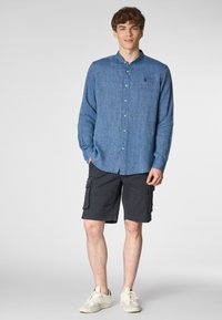 Camicia azzurra con colletto, maniche lunghe e una leggera texture; abbinata a pantaloni cargo neri e sneakers bianche.