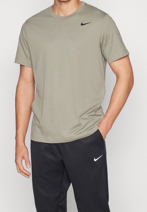 Homme portant un t-shirt Nike vert olive clair à manches courtes et un pantalon de survêtement Nike noir, les mains détendues dans la poche.
