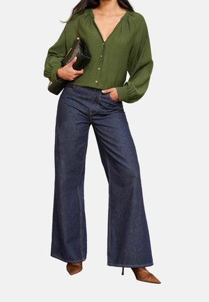 Kvinde iført en olivengrøn skjorte med knapper, højtaljede, vidde mørkeblå denimjeans, brune spidse hæle, holder en sort clutch.