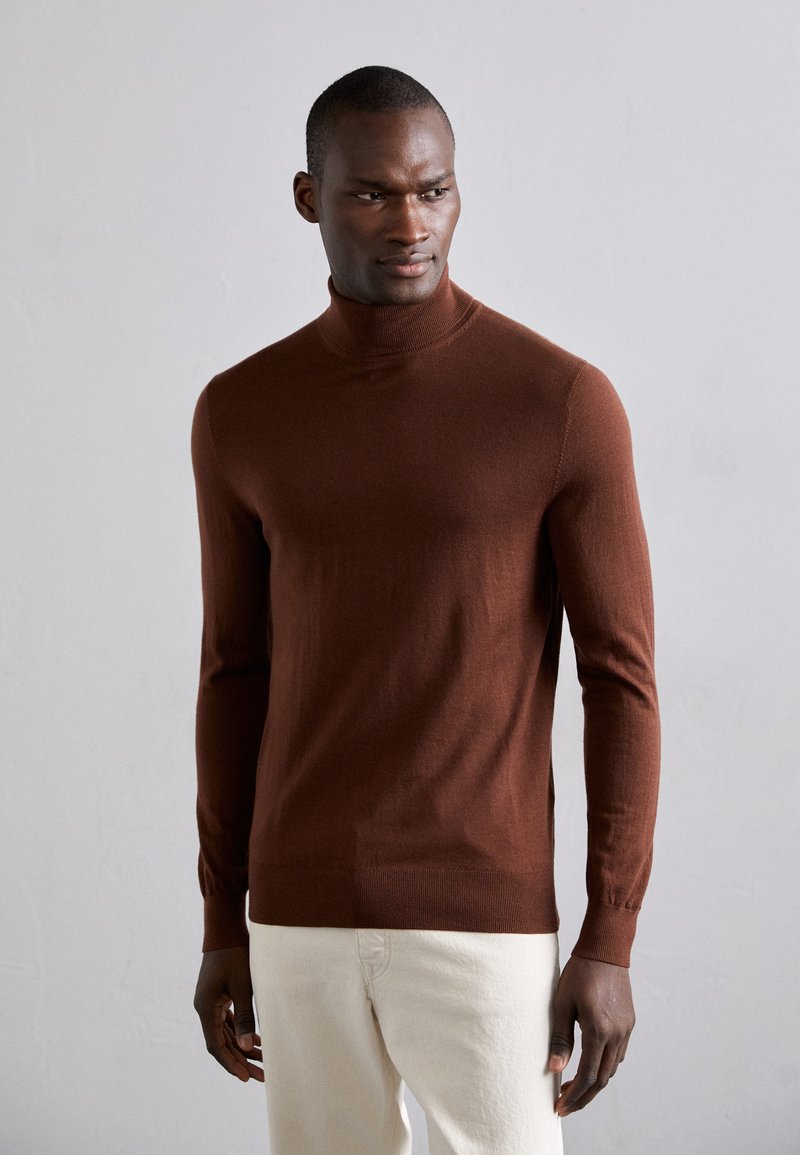 Rjava, dolga turtleneck pulover iz mehkega pletenega materiala, s prilegajočim se krojem in rebrastimi manšetami; v kombinaciji avec svetlimi hlačami.
