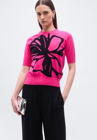 Γυναίκα που φοράει ένα φωτεινό ροζ κοντομάνικο τοπ με μεγάλο μαύρο λουλουδένιο μοτίβο, μαύρο παντελόνι και κρατάει μια ασημένια τσάντα φάκελο, μπροστά σε μονόχρωμο φόντο.