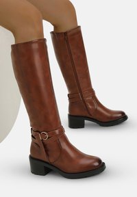 Botas marrones hasta la rodilla hechas de cuero sintético. Presentan un cierre de cremallera, un acento de hebilla y un tacón grueso. Textura suave en toda su superficie.