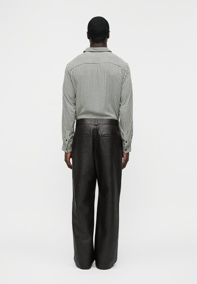 Homem em pé de costas, vestindo uma camisa branca de manga comprida aos quadrados, enfiada em calças pretas de perna larga e cintura alta.