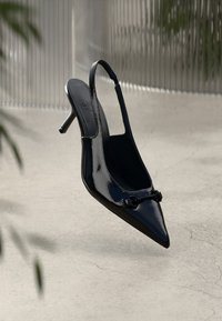 Salto slingback em cabedal envernizado preto com bico fino, salto fino e detalhe em nó decorativo na parte da frente. Textura lisa e brilhante.