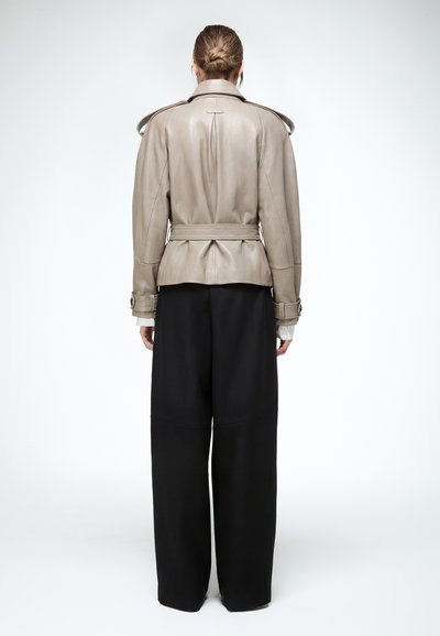 Haját kontyba fogó személy, bézs színű, övvel ellátott bőrkabátot és bő szárú, fekete nadrágot visel, háttal áll egy egyszínű háttér előtt.