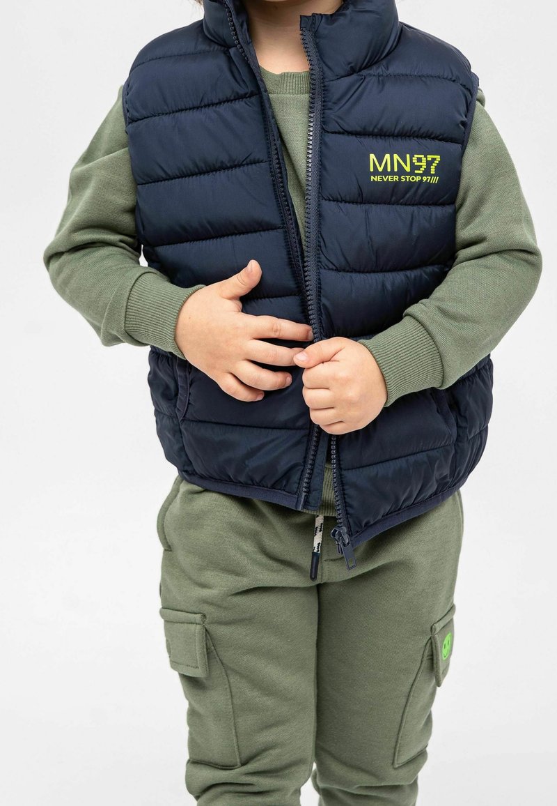 Enfant portant un sweat-shirt vert olive et un pantalon avec des poches cargo, attachant une veste matelassée bleu marine avec le logo jaune "MN97 Never Stop".