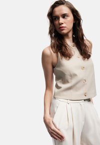 Beige ärmlös topp med hög halsringning och framknäppning, i kombination med ljusa plisserade byxor, mjukt tyg.