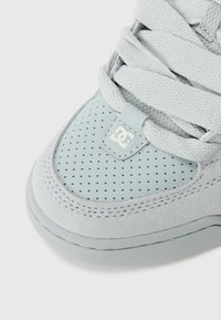 Ljusgrå sneaker med perforerad lädertå, mockasamling, tjocka texturerade snören och en liten DC-logga på snörhållaren.