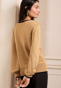 Un pull kaki avec un corps ajusté, de longues manches ballons et un bord noir festonné au col et à l'ourlet, porté sur un pantalon sombre.