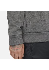 Gros plan sur une main tatouée portant une veste grise texturée avec l'inscription « THERMAL FIT » et un pantalon noir avec une poche zippée.