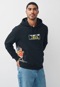 Zwarte hoodie met een Grinch-afbeelding op de borst en kleurrijke feestelijke ontwerpen op de zak. Gemaakt van zachte stof met een buidelzak.