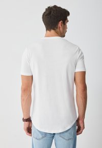 Camiseta blanca de manga corta con dobladillo redondeado, hecha de algodón. La parte trasera presenta un diseño simple y sin costuras, sin detalles adicionales.