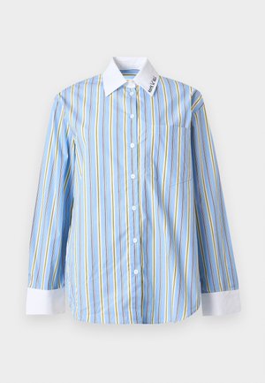 Frau trägt ein blau-weiß gestreiftes Hemd mit Knopfleiste, weißem Kragen und Manschetten, dunkle Jeans, schwarzen Gürtel und schwarze spitze Schuhe.