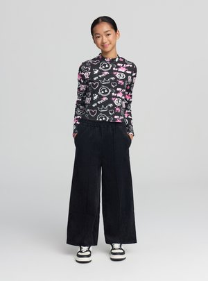 Giovane donna in piedi con le mani nelle tasche, indossa una maglia a maniche lunghe nera con motivo, pantaloni larghi neri e sneakers bianche e nere.