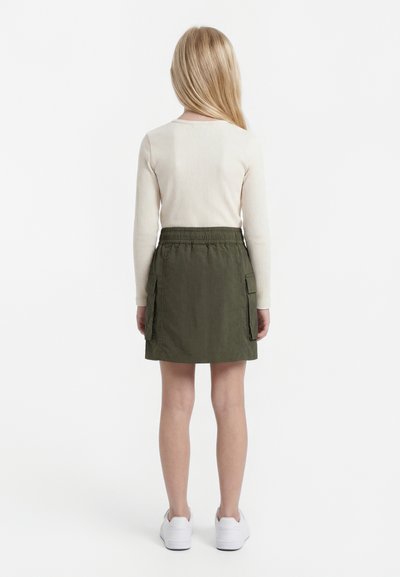 Vaikas su ilgais šviesiais plaukais stovi nugara, dėvi kreminės spalvos ilgomis rankovėmis palaidinę, alyvuogių žalios spalvos sijoną su kišenėmis ir baltus sportbačius.