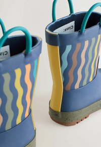 Bunte Gummistiefel mit gewellten Mustern in Blau, Gelb und Pastelltönen, mit türkisfarbenen Griffen und einer strukturierten braunen Außensohle.