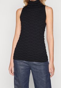 Czarny top bez rękawów z wysokim, marszczonym kołnierzem i teksturowanym geometrycznym wzorem. W połączeniu z niebieskimi dżinsami, prezentującymi przylegający krój.