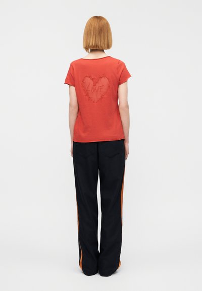 Μπλουζάκι σε χρώμα σκουριάς με σχέδιο καρδιάς και τη λέξη "LOVE" στο κέντρο, συνδυασμένο με μαύρο παντελόνι που έχει πορτοκαλί πλαϊνές ρίγες.