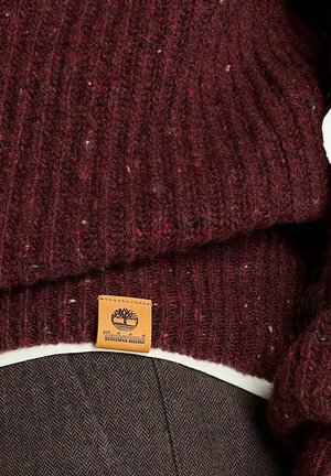 Maglione bordeaux a coste con una piccola etichetta tan del marchio Timberland sull'orlo, indossato sopra un capo bianco e pantaloni marroni.