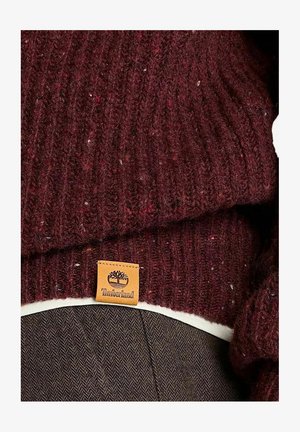 Maglione bordeaux a coste con una piccola etichetta tan del marchio Timberland sull'orlo, indossato sopra un capo bianco e pantaloni marroni.