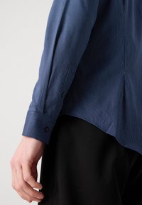 Cămașă cu dungi de culoare bleumarin, cu mansete cu buton, având dungi fine albe, textură netedă și un design cu tiv drept.